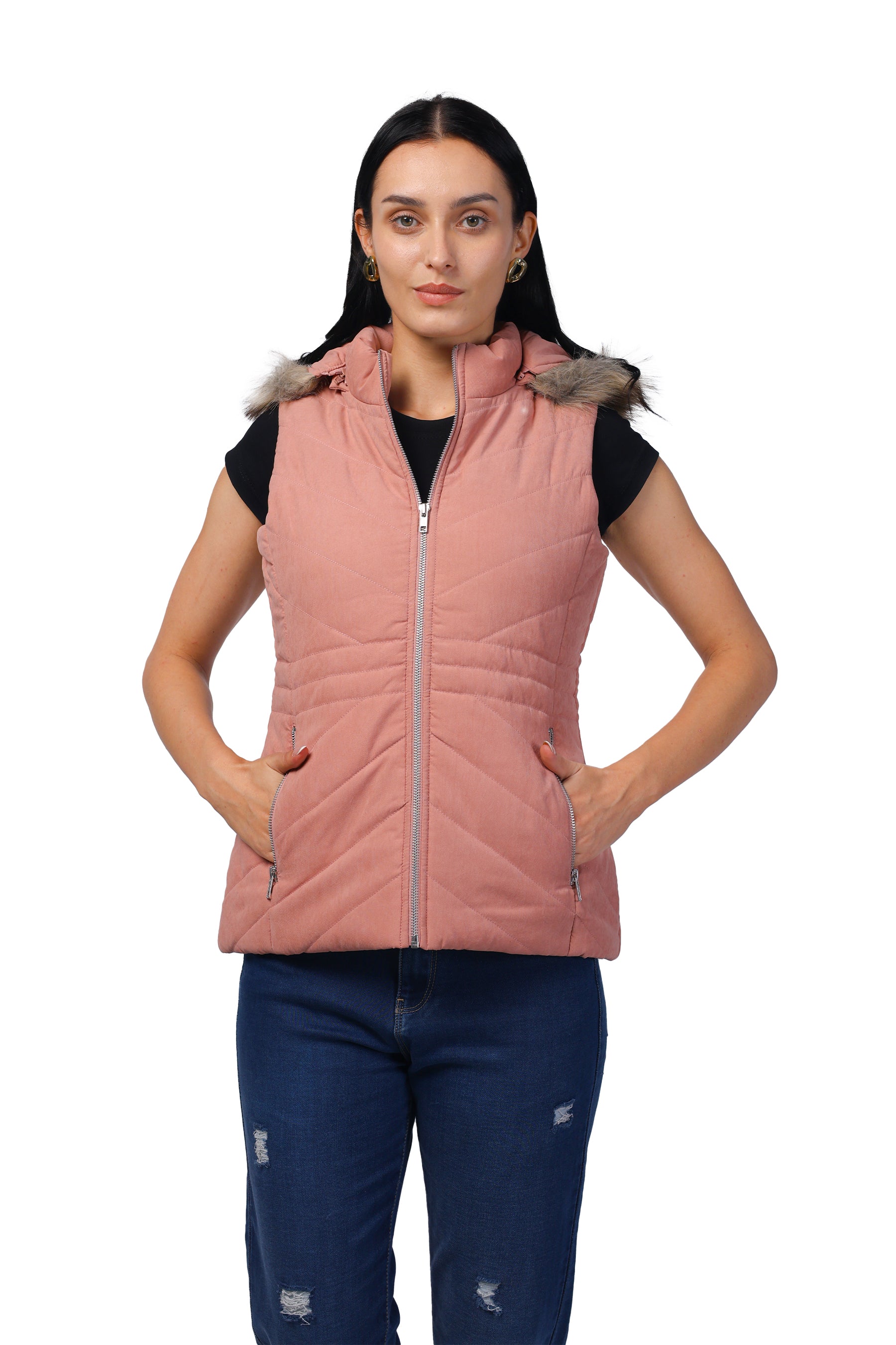 A woman modeling a sleeveless quilted puffer vest in dusty pink, featuring a faux-fur trimmed hood and a front zipper, paired with a black top and blue jeans against a plain white background.