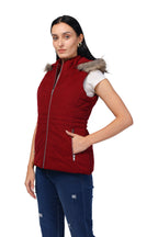 A woman modeling a sleeveless burgundy puffer vest with a gray faux-fur trimmed hood, worn over a white t-shirt and blue jeans, against a plain white background.