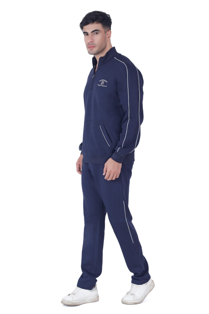 A man in coatsnmore’s navy blue half-zip tracksuit with white piping and a chest logo, standing sideways in a casual pose, wearing white sneakers against a white background.