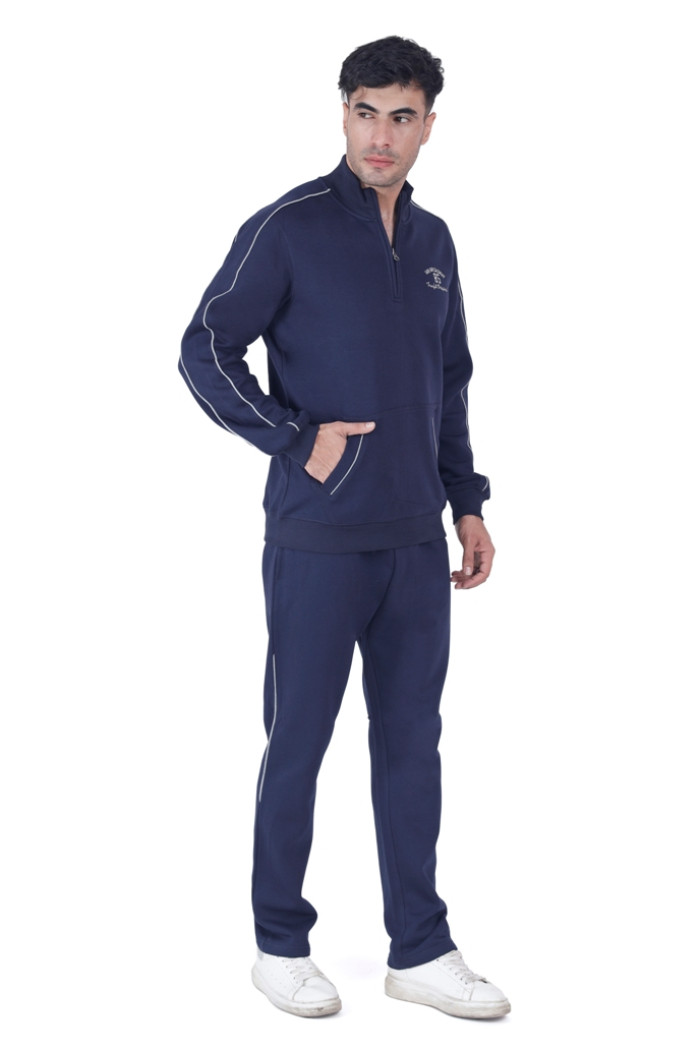A man wearing coatsnmore’s navy blue tracksuit with a half-zip design, white piping, and chest branding, standing in a relaxed pose with one hand in the pocket, paired with white sneakers against a plain white background.