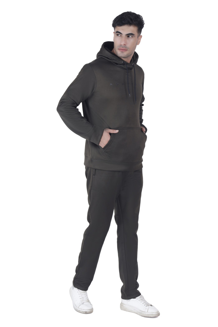 A man wearing Coatsnmore’s olive hoodie-style tracksuit with a kangaroo pocket and drawstrings, standing with both hands in the pocket, paired with white sneakers against a plain white background.