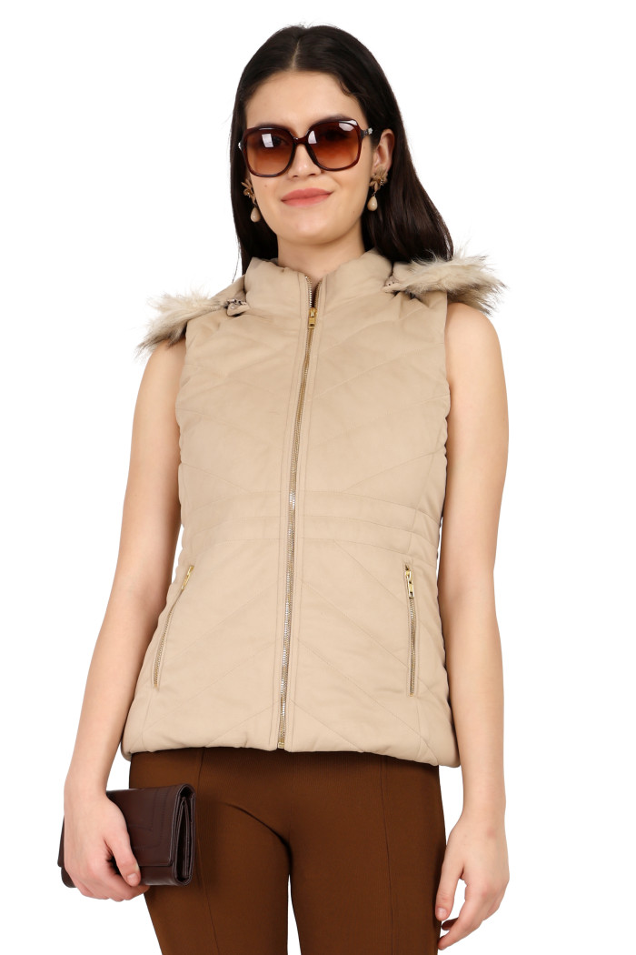 A standing side pose of a woman wearing Coatsnmore’s half sleeves jacket in beige colour with a textured pattern, removable hood with faux fur trim, side zippered pockets, a zip closure and coffee jeans holding a clutch from her right hand.