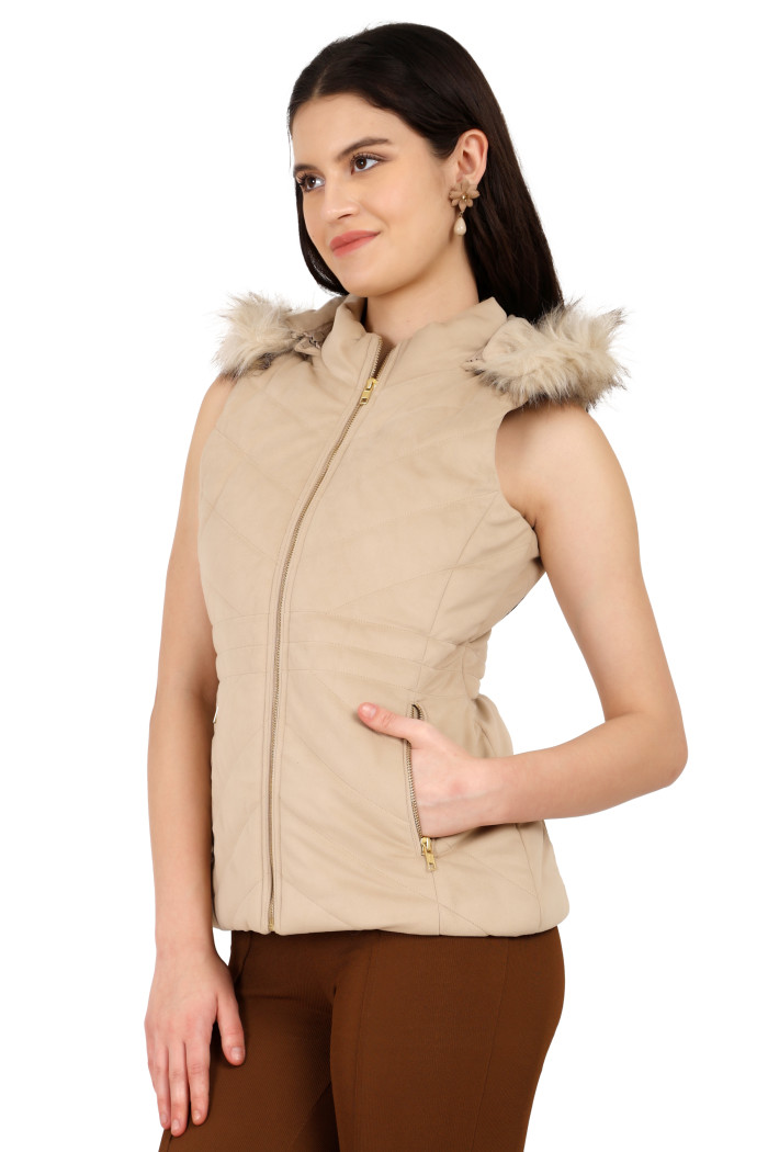 A standing side pose of a woman wearing Coatsnmore’s half sleeves jacket in beige colour with a textured pattern, removable hood with faux fur trim, side zippered pockets, a zip closure and coffee jeans with her left hand in the pocket.