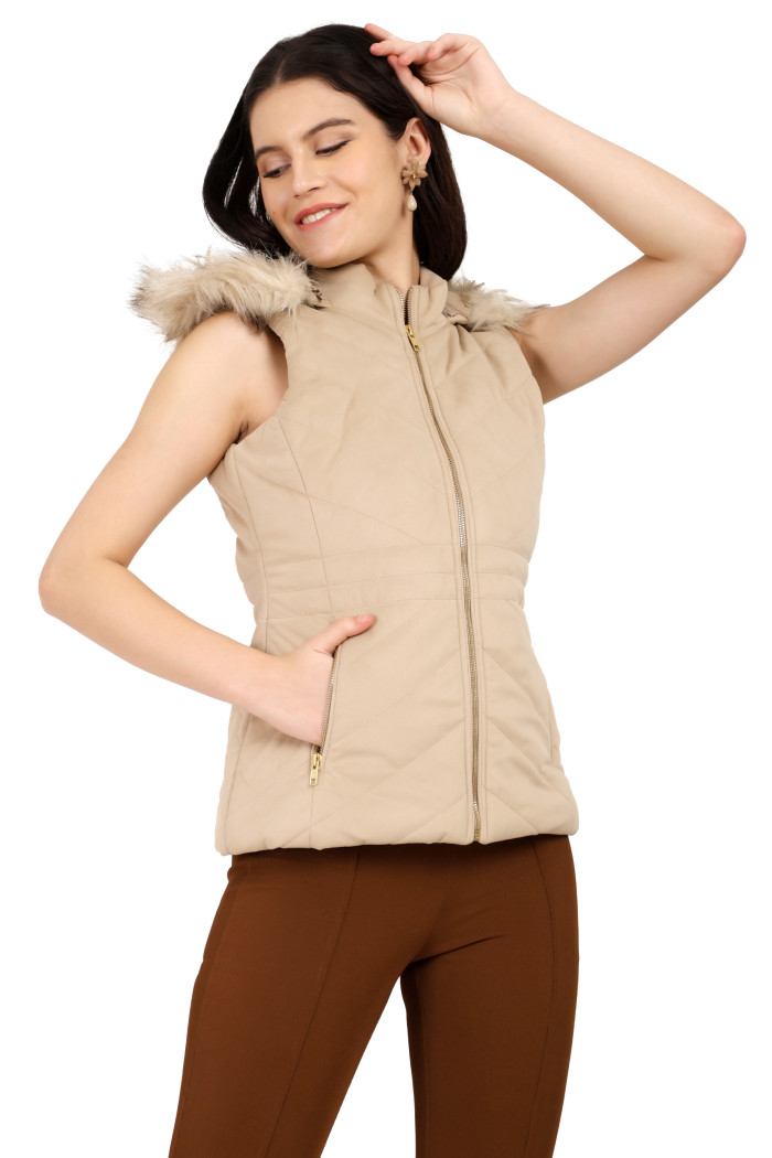 A standing side pose of a woman wearing Coatsnmore’s half sleeves jacket in beige colour with removable hood with faux fur trim, side zippered pockets, a zip closure and coffee jeans with her right hand in pocket and left hand on her head.