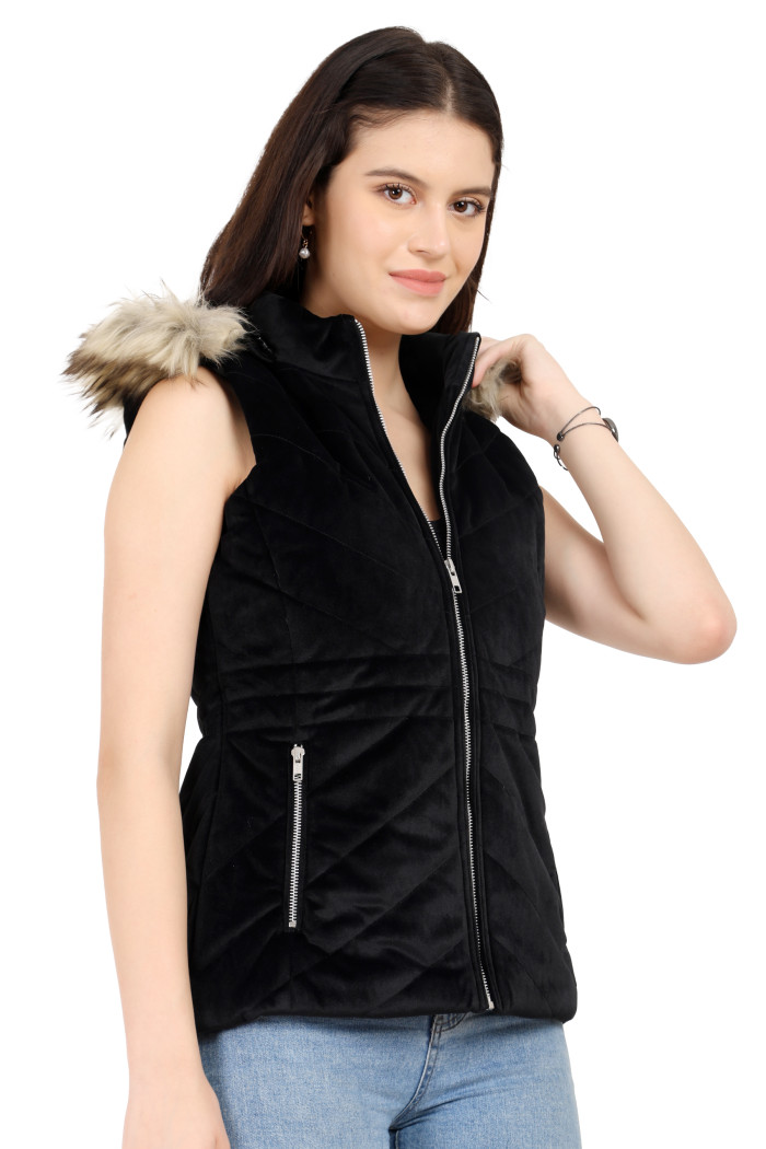 A standing side pose of a woman wearing Coatsnmore’s half sleeves jacket in black colour with a textured pattern, removable hood with faux fur trim, side zippered pockets, a zip closure and blue jeans holding the hood.