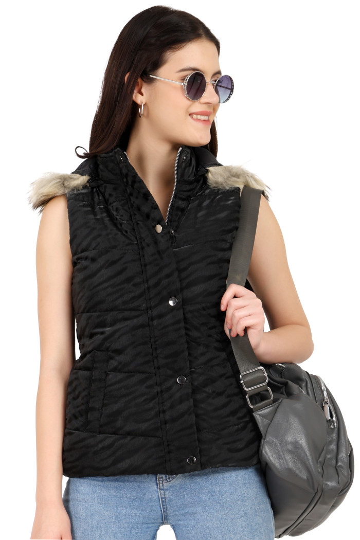 A standing pose of a woman wearing shades, Coatsnmore’s black quilted jacket with a high neck, zip closure, removable hood with faux fur trim, button plackets, and blue jeans, holding a bag.