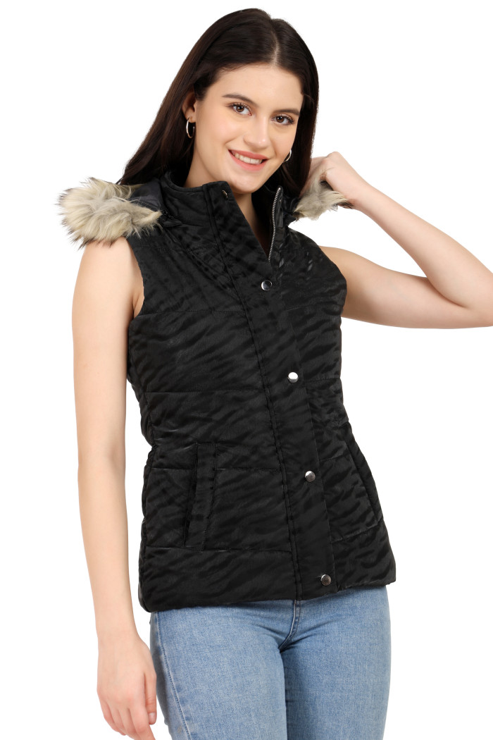 A standing pose of a woman wearing Coatsnmore’s black quilted jacket with a high neck, zip closure, removable hood with faux fur trim, button plackets, and blue jeans, holding the hood with her left hand.