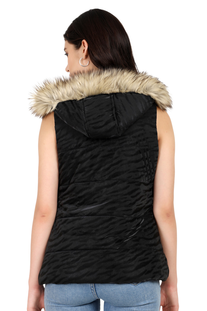 A back pose of a standing woman, wearing Coatsnmore’s black quilted jacket with a high neck, zip closure, removable hood with faux fur trim, button plackets, and blue jeans.