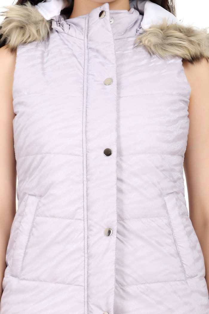 A standing pose of a woman wearing Coatsnmore’s lilac sleeveless quilted jacket with a zip closure, mandarin collar, removable hood, button plackets and black jeans with her right hand in the pocket.