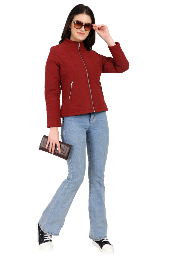 A woman in a standing pose, wearing Trufit’s maroon quilted biker jacket with a Chinese collar, sliver zip closure, and side pockets with a silver zipper and blue jeans, holding black shades with her left hand and a clutch in her right hand.