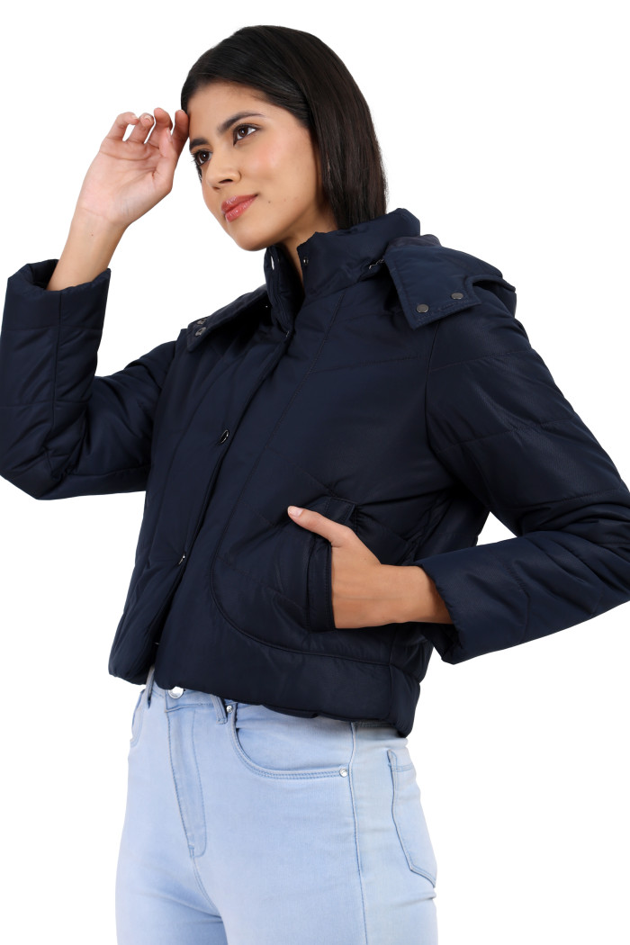 A woman in a standing side pose, wearing Coatsnmore’s black quilted crop jacket featuring a Chinese collar, removable hood, zip closure with button closure, side pockets and blue jeans with her right hand on head and left hand in the pocket.