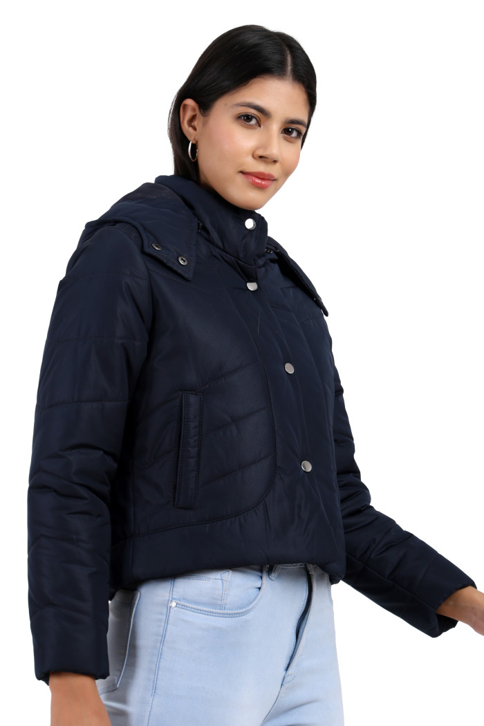 A woman is giving a standing side pose, wearing Coatsnmore’s navy quilted loose-fit crop jacket featuring a Chinese collar, a removable hood, a zip closure with button closure, side pockets and blue jeans.