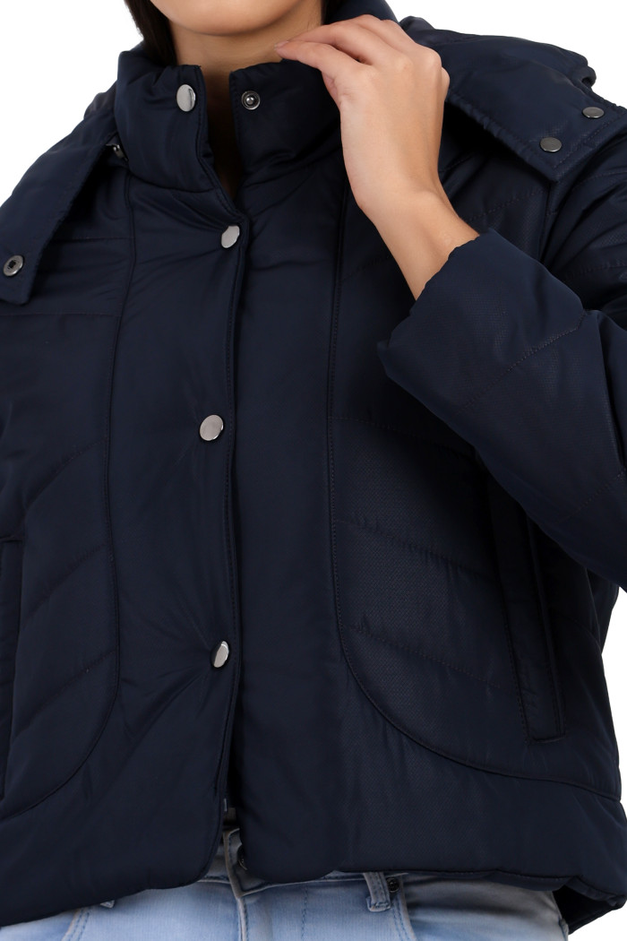 A woman in a cropped image is wearing Coatsnmore’s navy quilted loose-fit crop jacket featuring a Chinese collar, a removable hood, a zip closure with button closure and side pockets holding her collar.