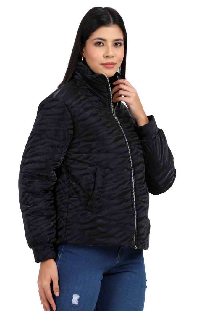 A woman in a standing side pose, wearing Trufit’s black quilted jacket with a high neck collar, zip closure, side pockets, blue jeans and holding the jacket’s collar with her right hand.