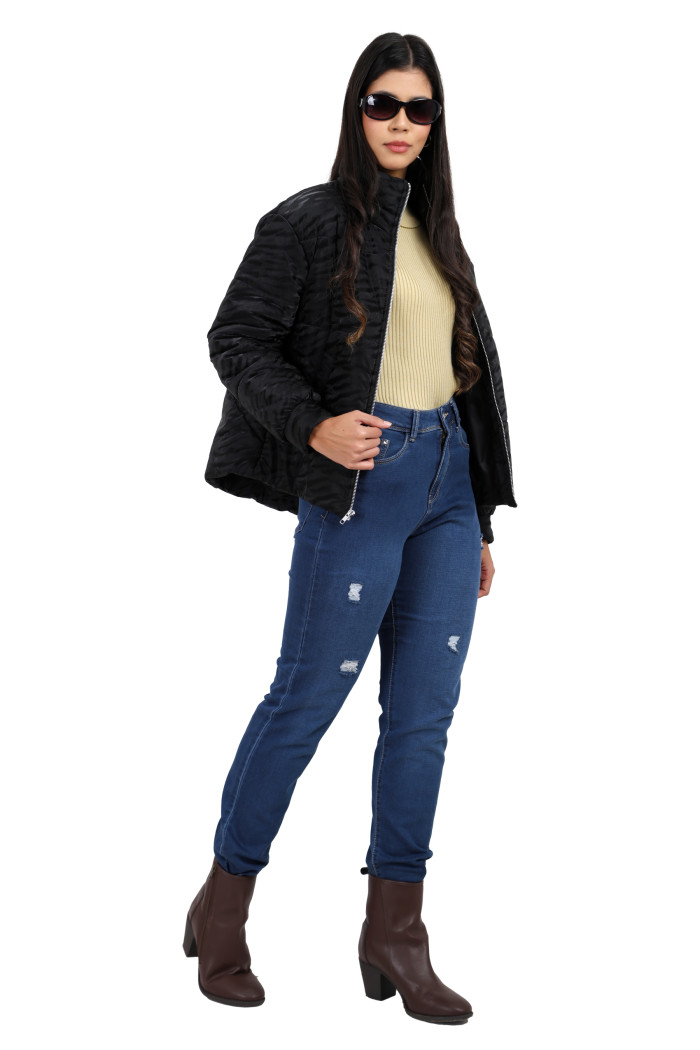 A woman in a standing pose in black shades, wearing Trufit’s black quilted jacket with a high neck, zip closure, side pockets, and blue jeans, is holding the jacket with her right hand.