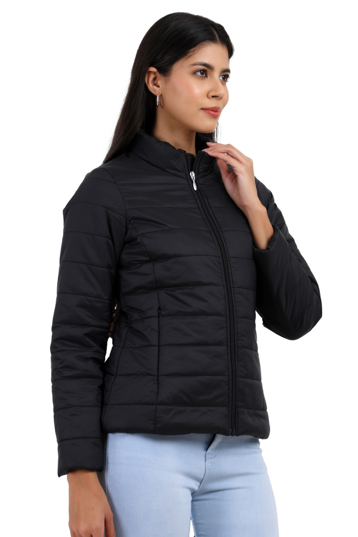 A side profile of a woman standing highlights Coatsnmore’s black quilted jacket with its high neck collar, sleek zip closure, hidden side pockets, and blue jeans.