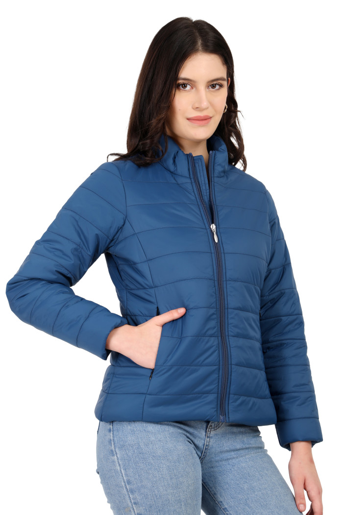 A side profile of a woman standing highlights Coatsnmore’s blue quilted jacket with its high neck collar, sleek zip closure, hidden side pockets, and blue jeans.