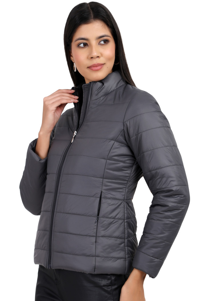 A woman in a standing side pose is wearing Coatsnmore’s grey quilted jacket with a high neck collar, zip closure, side pockets, and black jeans while holding her collar with her right.