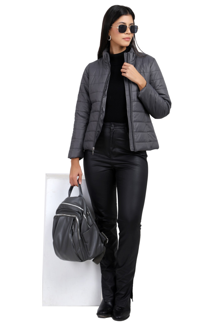 A woman in a standing pose, wearing Coatsnmore’s grey quilted jacket with a high neck, zip closure, side pockets, and black jeans, is holding a bag with her right hand and holding her jacket with her left hand.