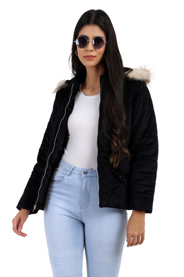 A woman is giving a standing pose in black shades, wearing Trufit’s black velvet quilted jacket with a a zip closure, removable hood with faux fur trim, high neck, slash pockets and blue jeans.