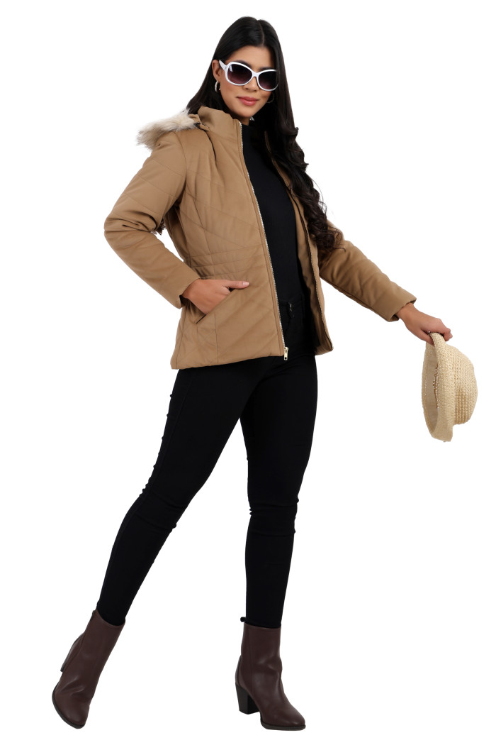 A woman in a standing pose in black shades, wearing Coatsnmore’s coffee velvet quilted jacket with a high neck, zip closure, removable hood with faux fur trim, slash pockets, and blue jeans, while holding her jacket from her left hand.