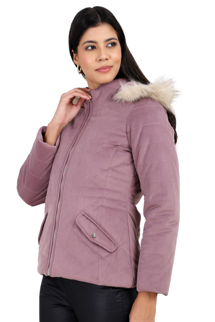 A woman in a standing side pose is wearing Coatsnmore’s lilace velvet quilted jacket with a high neck, zip closure, removable hood with faux fur trim, slash pockets, and black jeans while holding her jacket’s collar with her left hand.