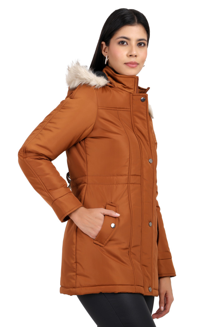 A woman is giving a standing side pose, wearing Coatsnmore’s rust quilted parka jacket with a detachable hood, zip closure, button placket, side pockets and quilted pattern and black jeans with her right hand in pocket.