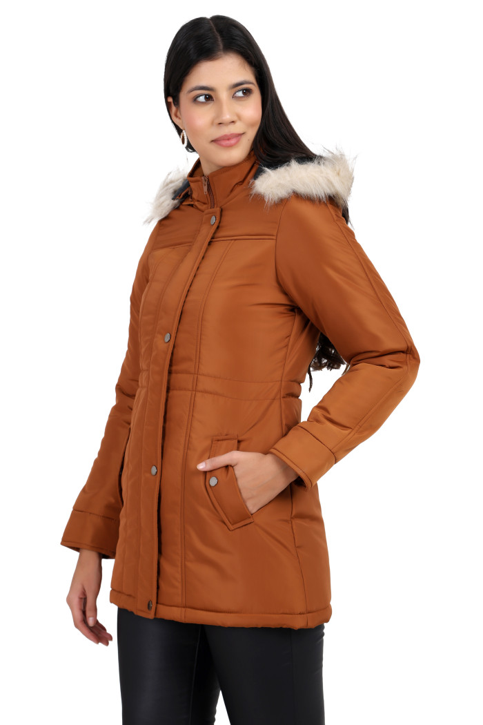 A woman is giving a standing left side pose, wearing Coatsnmore’s rust quilted parka jacket with a detachable hood, zip closure, button placket, side pockets and quilted pattern and black jeans with left hand in pocket.