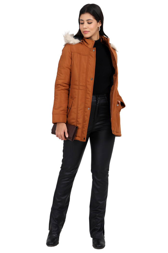 A woman is giving a standing pose, wearing Coatsnmore’s rust quilted parka jacket with a detachable hood, zip closure, button placket, side pockets and quilted pattern, black jeans and holding the clutch from her right hand and right hand in pocket.