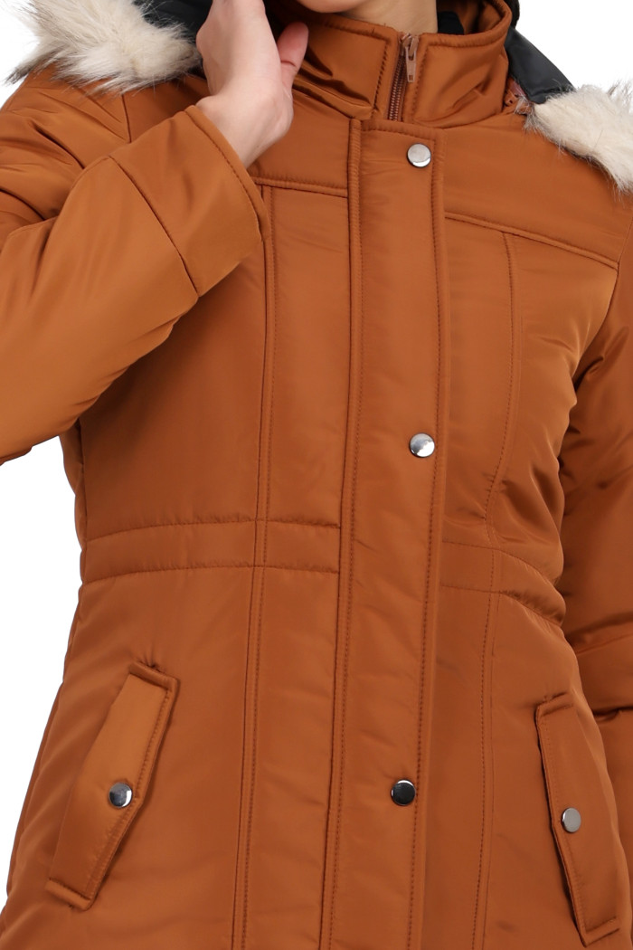 A woman in a cropped image is wearing Coatsnmore’s rust quilted parka jacket with a detachable hood, zip closure, button placket, side pockets and quilted pattern with her right hand on her neck.