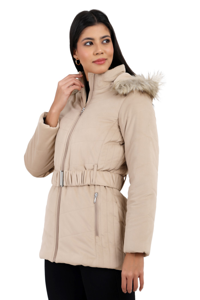 A woman in a side pose showcases Coatsnmore’s beige velvet quilted jacket, featuring a mandarin collar, removable hood, adjustable waistband zip closure, concealed zippered side pockets, and black jeans with her right hand on the collar.