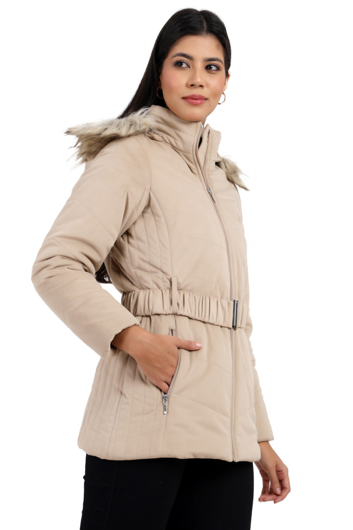 A side profile of a woman standing highlights Coatsnmore’s beige velvet quilted jacket, featuring a mandarin collar, removable hood, adjustable waistband zip closure, concealed zippered side pockets and black jeans with her right hand in a pocket.