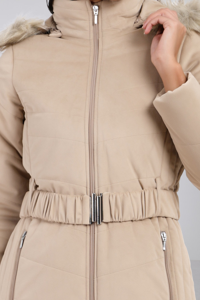 A cropped view of a woman in Coatsnmore’s beige velvet quilted jacket, featuring a mandarin collar, removable hood, adjustable waistband zip closure, and concealed zippered side pockets, holding the hood with her left hand.