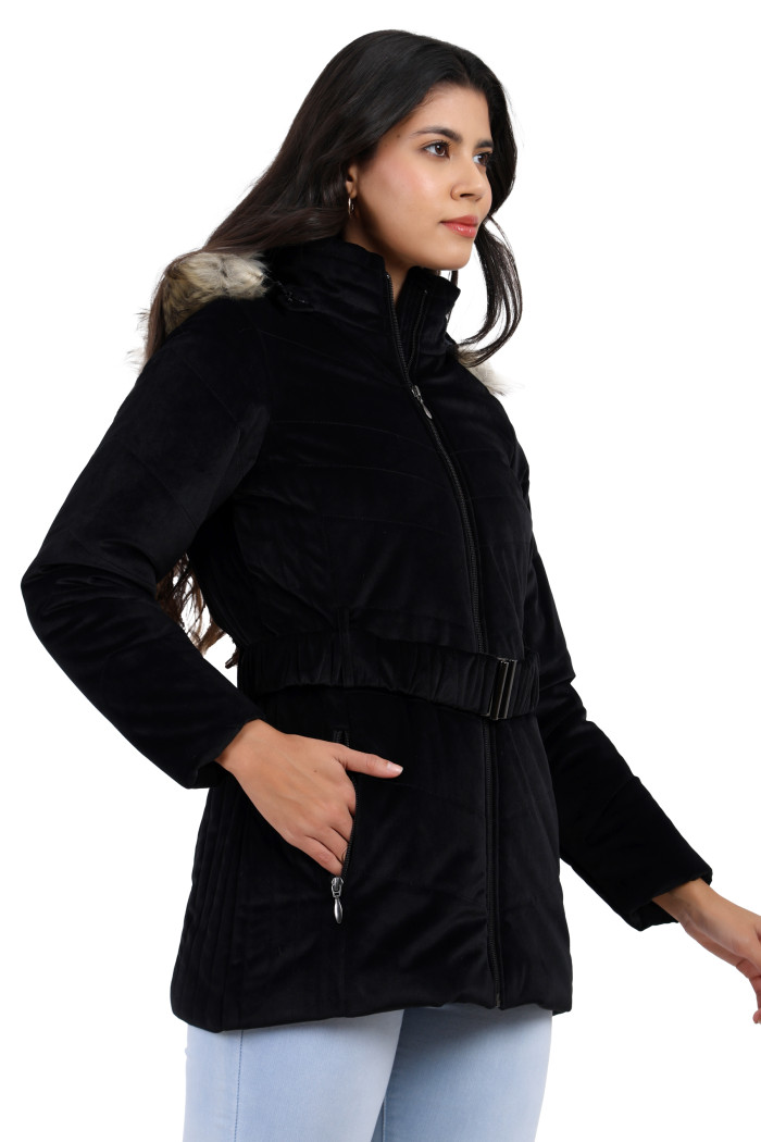 A woman in a side pose showcases the Coatsnmore’s black velvet quilted jacket, featuring a mandarin collar, removable hood, adjustable waistband zip closure, concealed zippered side pockets, and blue jeans with her right hand in the pocket.