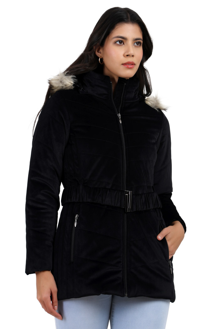 A side profile of a woman standing highlights Coatsnmore’s black velvet quilted jacket, featuring a mandarin collar, removable hood, adjustable waistband zip closure, concealed zippered side pockets and blue jeans with her left hand in a pocket.