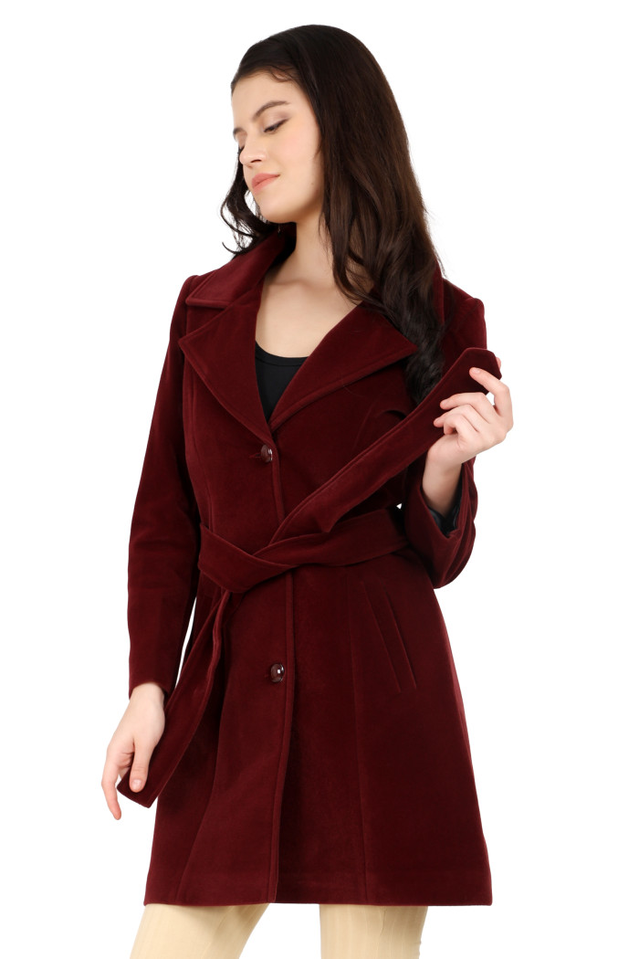 A woman is giving a standing side pose, wearing Trufit’s Long wine Velvet coat with a lapel collar, button closure, belt, side pockets, and skin bell bottom and holding the coat’s belt.