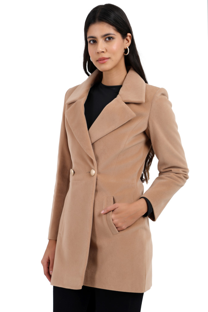 A woman is giving a standing side pose wearing Trufit’s double-breasted velvet coat in camel colour with a lapel collar, side pockets, button closure, and black jeans with her left hand in the pocket.