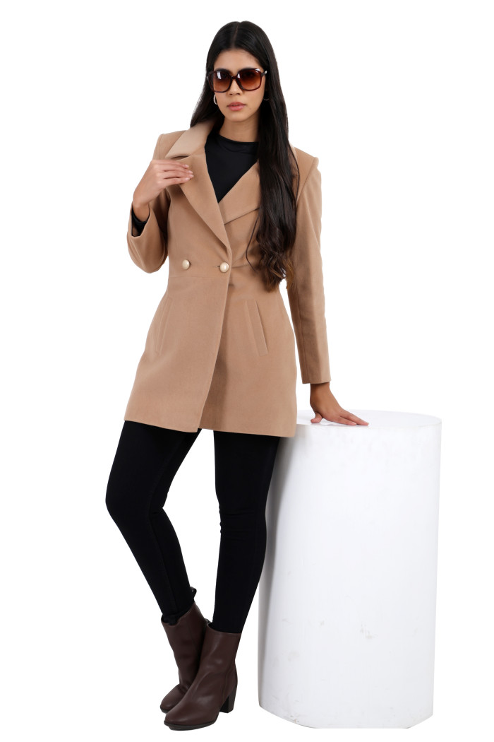 A woman in a standing pose is wearing black shades, Trufit’s double-breasted velvet coat in camel colour with a lapel collar, side pockets, button closure, and black jeans while holding the collar with her right hand.