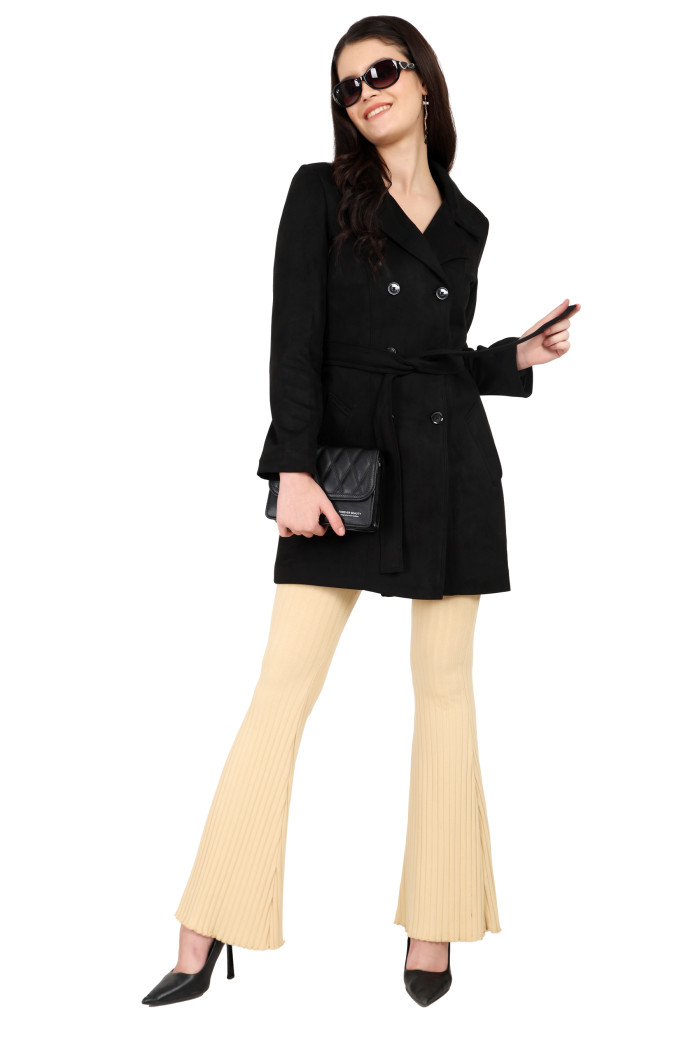 A woman is giving a standing pose in black shades, wearing Coatsnmore’s black double-breasted trench coat with a lapel collar, button closure, a belt, side pockets and skin bell bottoms and holding a clutch from her right hand and holding the coat’s belt.