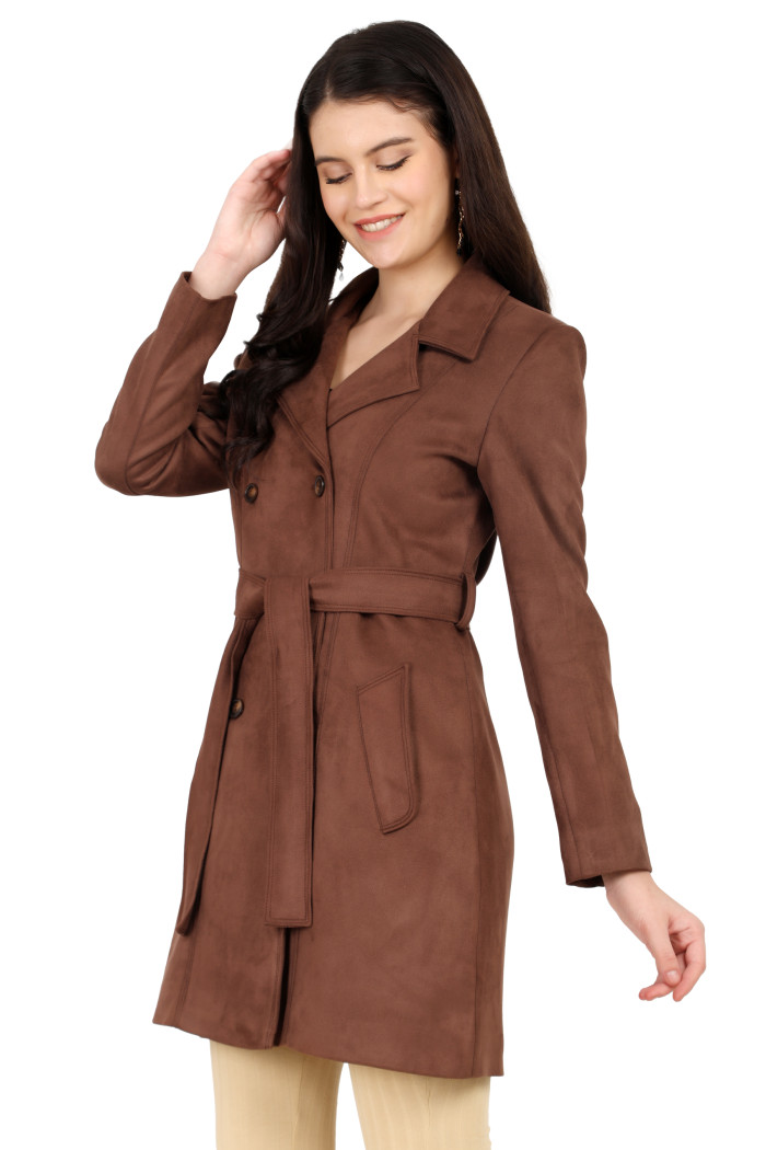 A woman is holding a standing side pose, wearing Coatsnmore’s coffee double-breast trench coat with a lapel collar, button closure, belt, side pockets, and skin bell bottom and touching her hair from her right hand.