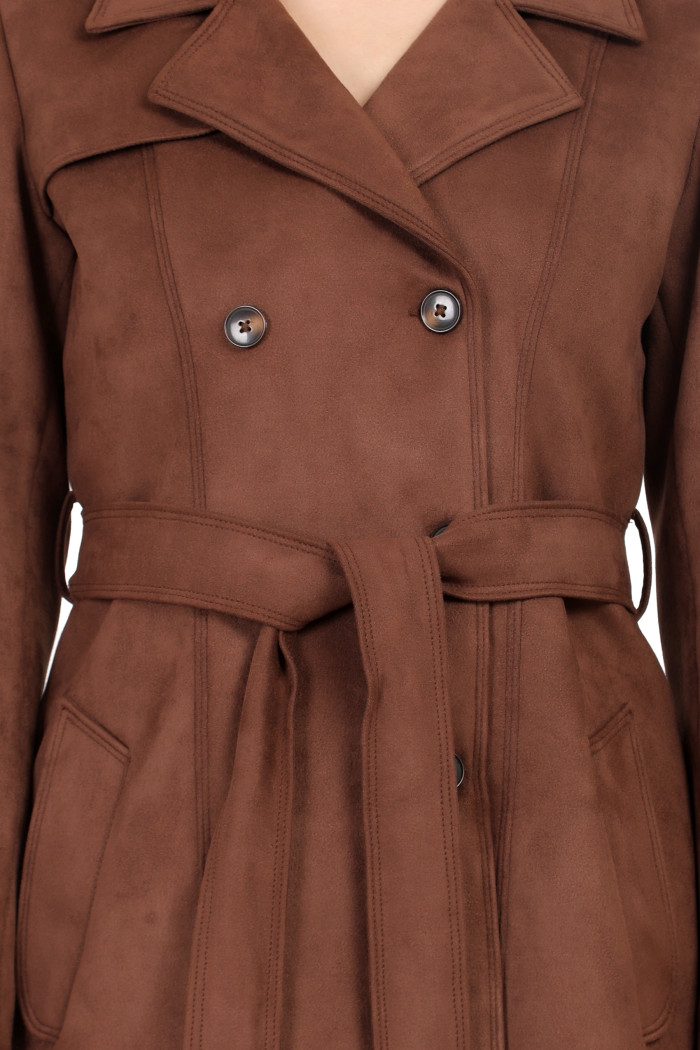 A woman in a cropped image is wearing Coatsnmore’s coffee double-breast trench coat with a lapel collar, button closure, belt and side pockets.