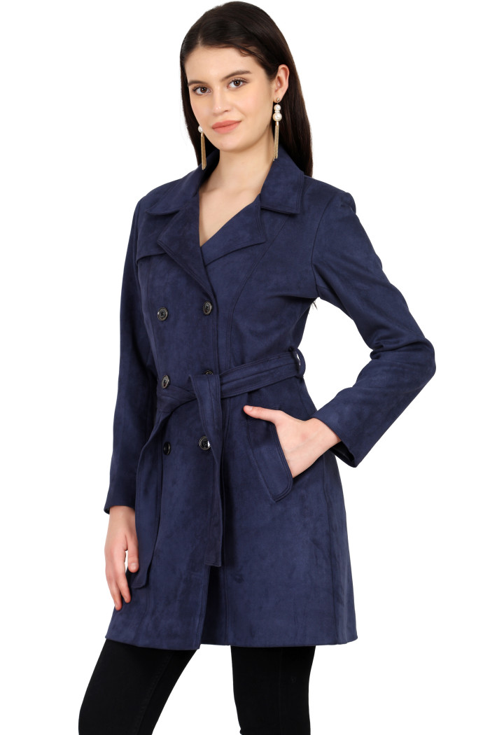 A woman is holding a standing side pose, wearing Coatsnmore’s navy double-breasted trench coat with a lapel collar, button closure, belt, side pockets, and black bottom with her left hand in the pocket.