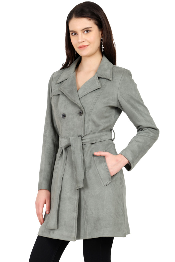 A woman is holding a standing side pose, wearing Coatsnmore’s pista double-breast trench coat with a lapel collar, button closure, belt, side pockets, and black bottom with her left hand in the pocket.