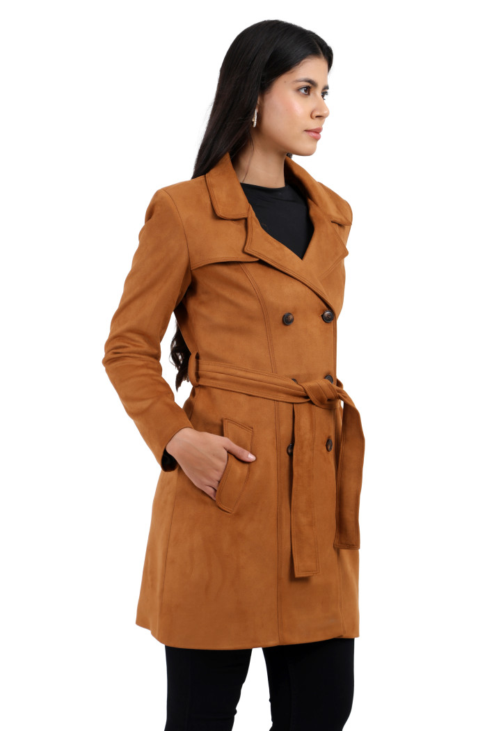 A woman is holding a standing side pose, wearing Coatsnmore’s tan double-breasted trench coat with a lapel collar, button closure, belt, side pockets, and black bottom with her right hand in the pocket.