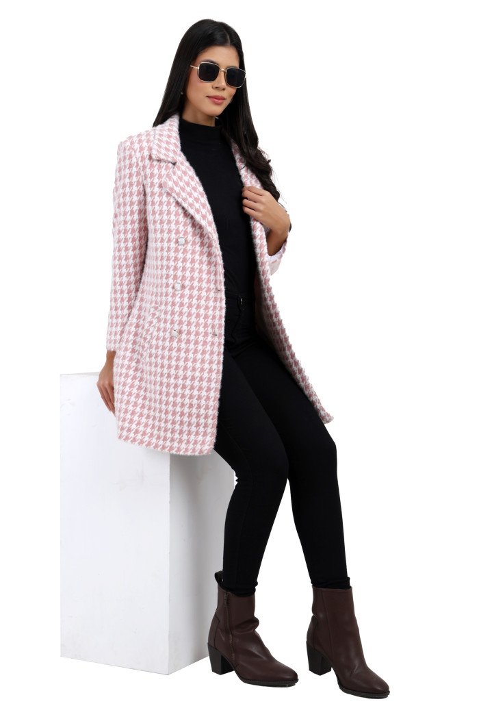 A woman sits elegantly in Coatsnmore’s pink double-breasted semi-long coat featuring a lapel collar, button-up design, side pockets, styled in black jeans while holding the collar from her left hand.