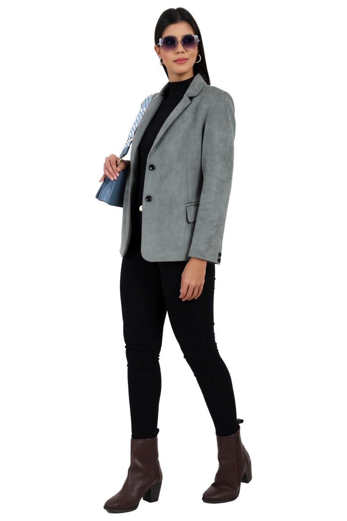 A woman in a standing side pose is wearing Trufit’s short-length coat in pista with a lapel collar, side pockets and a button closure in black jeans while holding the clutch in her right hand.